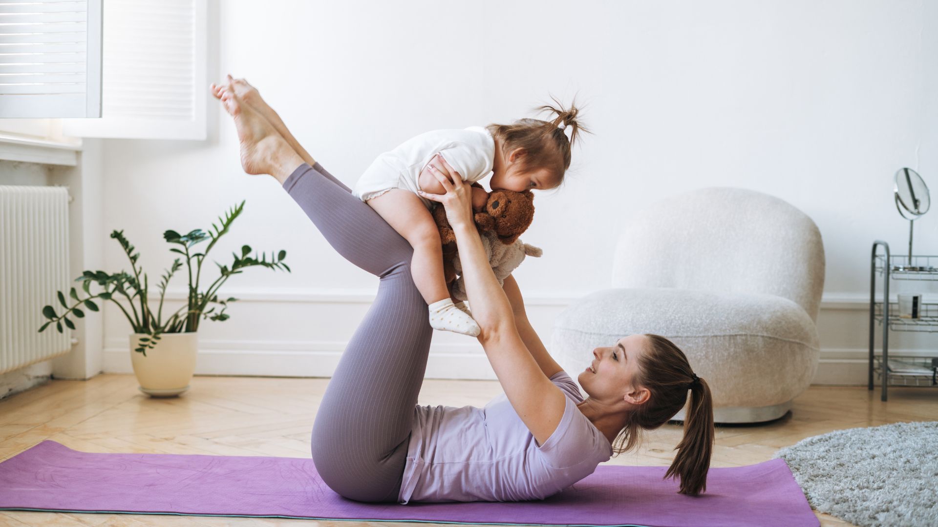 Imagem de uma sala com poltrona branca e um vaso de planta. Em destaque mãe e filha fazendo alogamento. A mãe segura a filha em seus braços, apoiando-a em suas pernas. A mãe está deitada sobre  um tapete lilás.