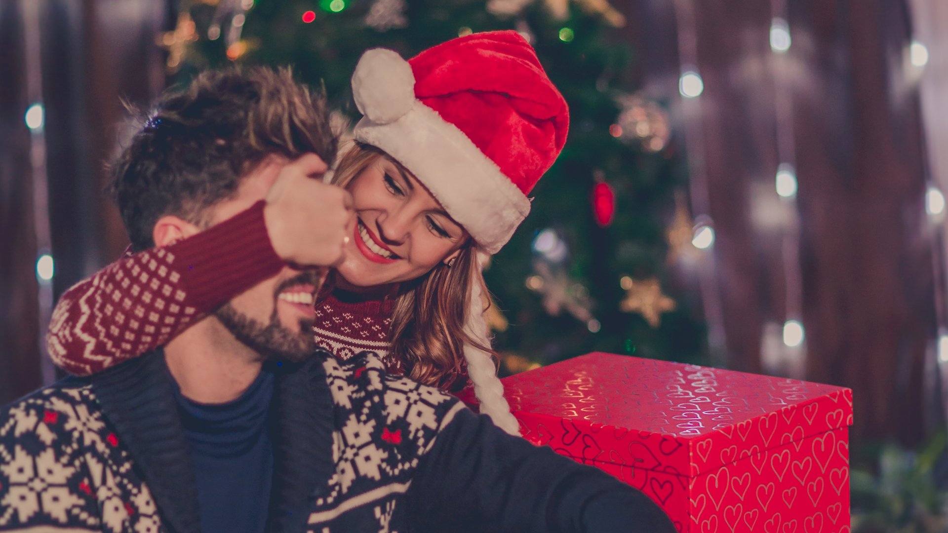 StefaNikolic / Getty Images Signature / Canva Casal no natal, no qual a mulher está cobrindo os olhos do homem para fazer uma surpresa
