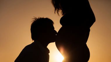 Homem beijando barriga da esposa grávida. No fundo, o sol está se pondo