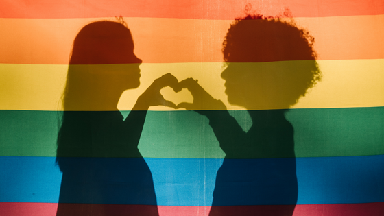 Silhueta de duas mulheres fazendo um coração com as mãos e ao fundo, a bandeira do orgulho LGBTQIAP+