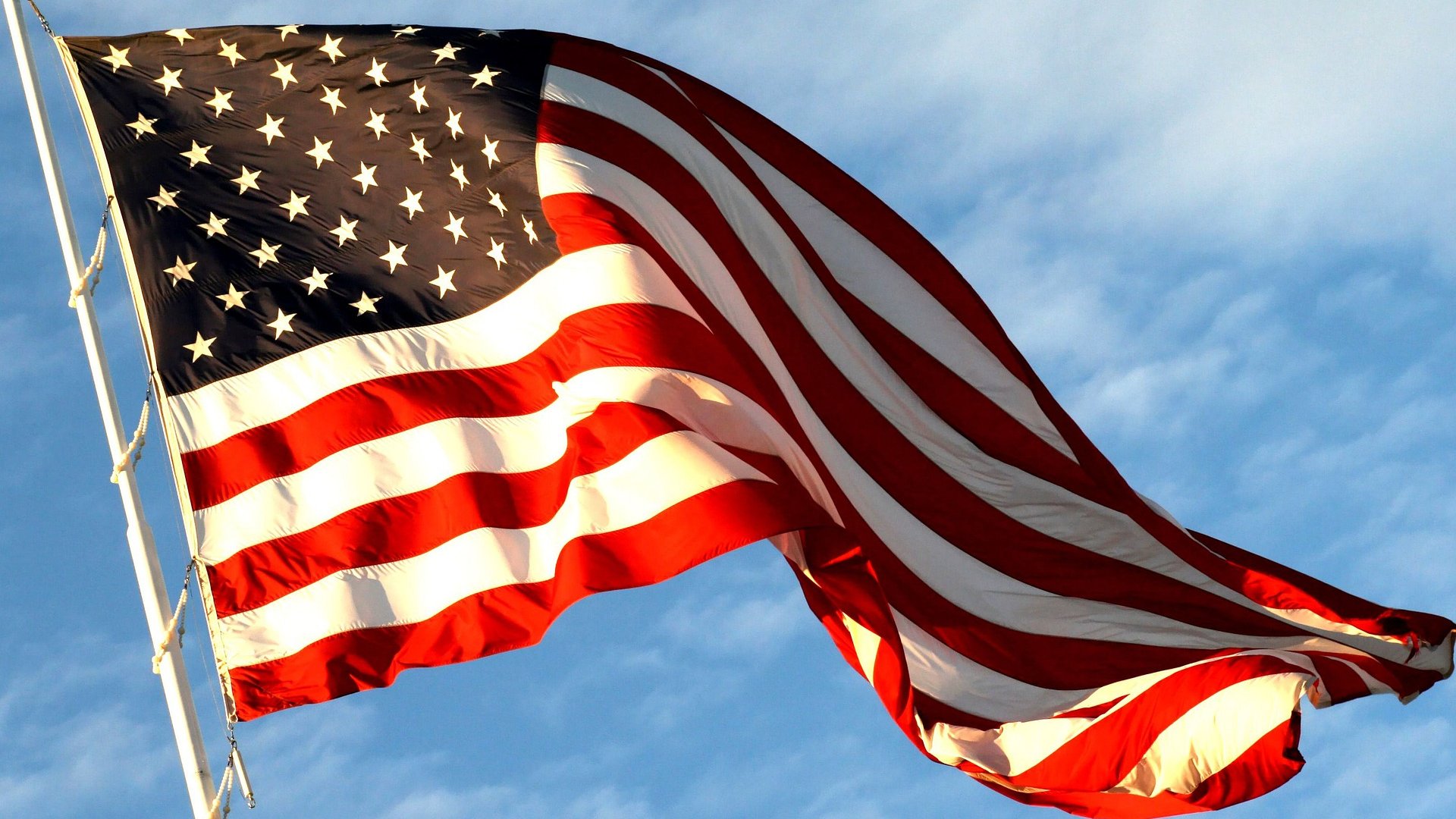 Bandeira dos Estados Unidos sendo mexida com o vento. Está pendurada em uma haste e o céu está ensolarado