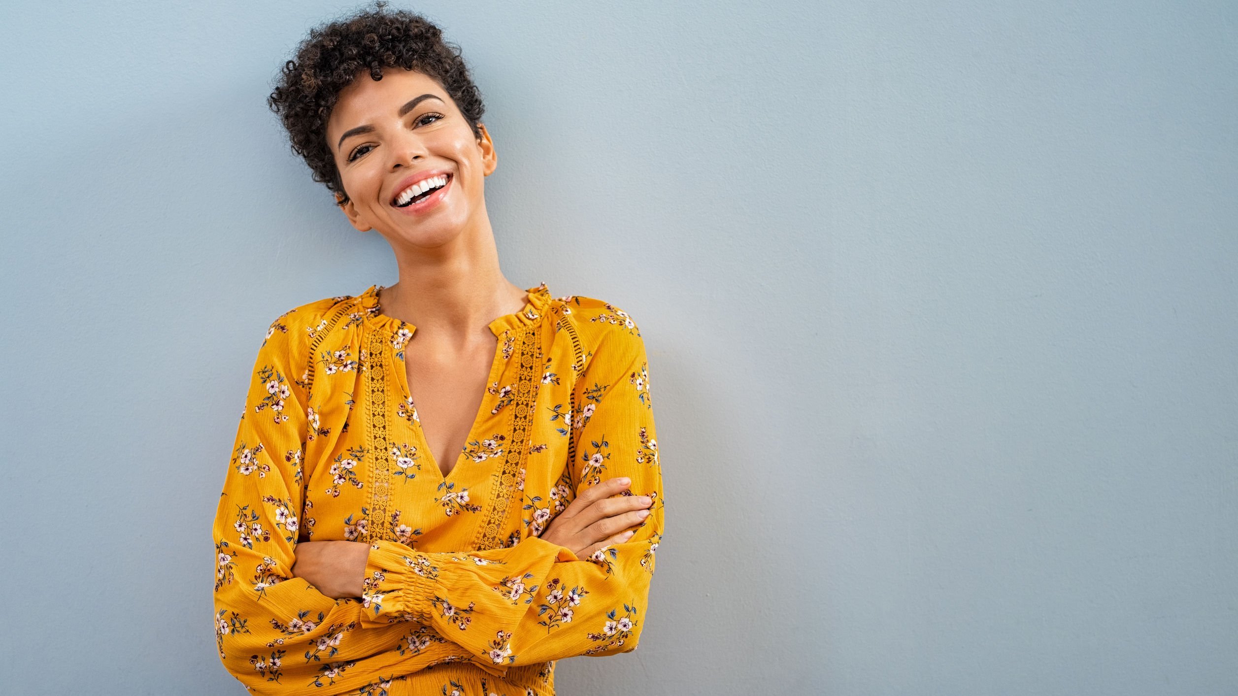 Mulher encostada na parede sorrindo com os braços cruzados