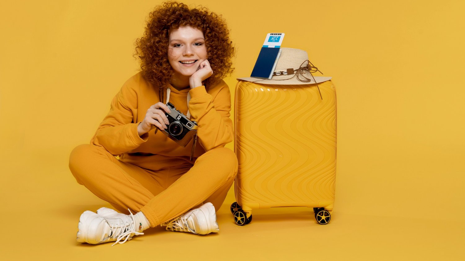 Mulher sorridente sentada no chão, ao lado de sua mala de viagem, segurando uma câmera antiga na mão