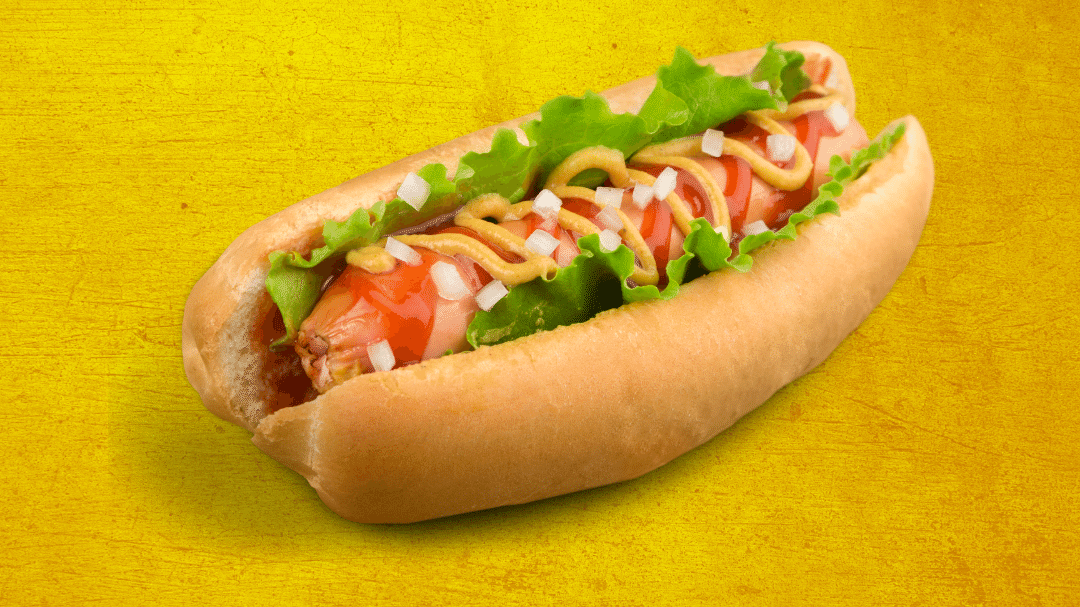 Imagem de um cachorro quente, com cebola, salada de alface, ketchup e mostarda. A cor da imagem de fundo é amarela.
