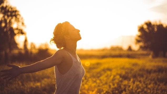 Mulher com os braços aberto, respirando, com o sol raiando atrás