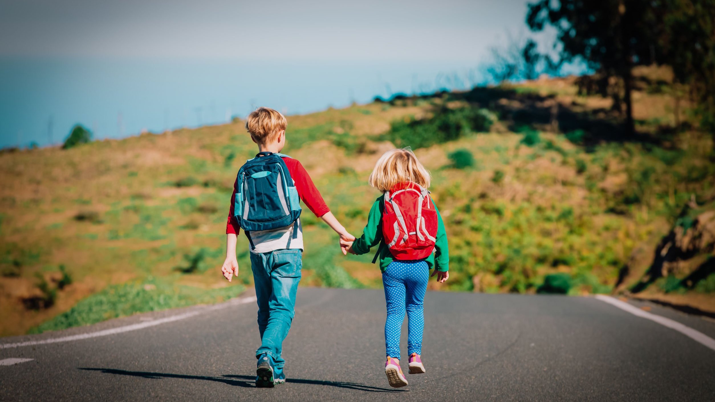 Menino e menina brancos e loiros andando de mãos dadas numa estrada com mochila nas costas