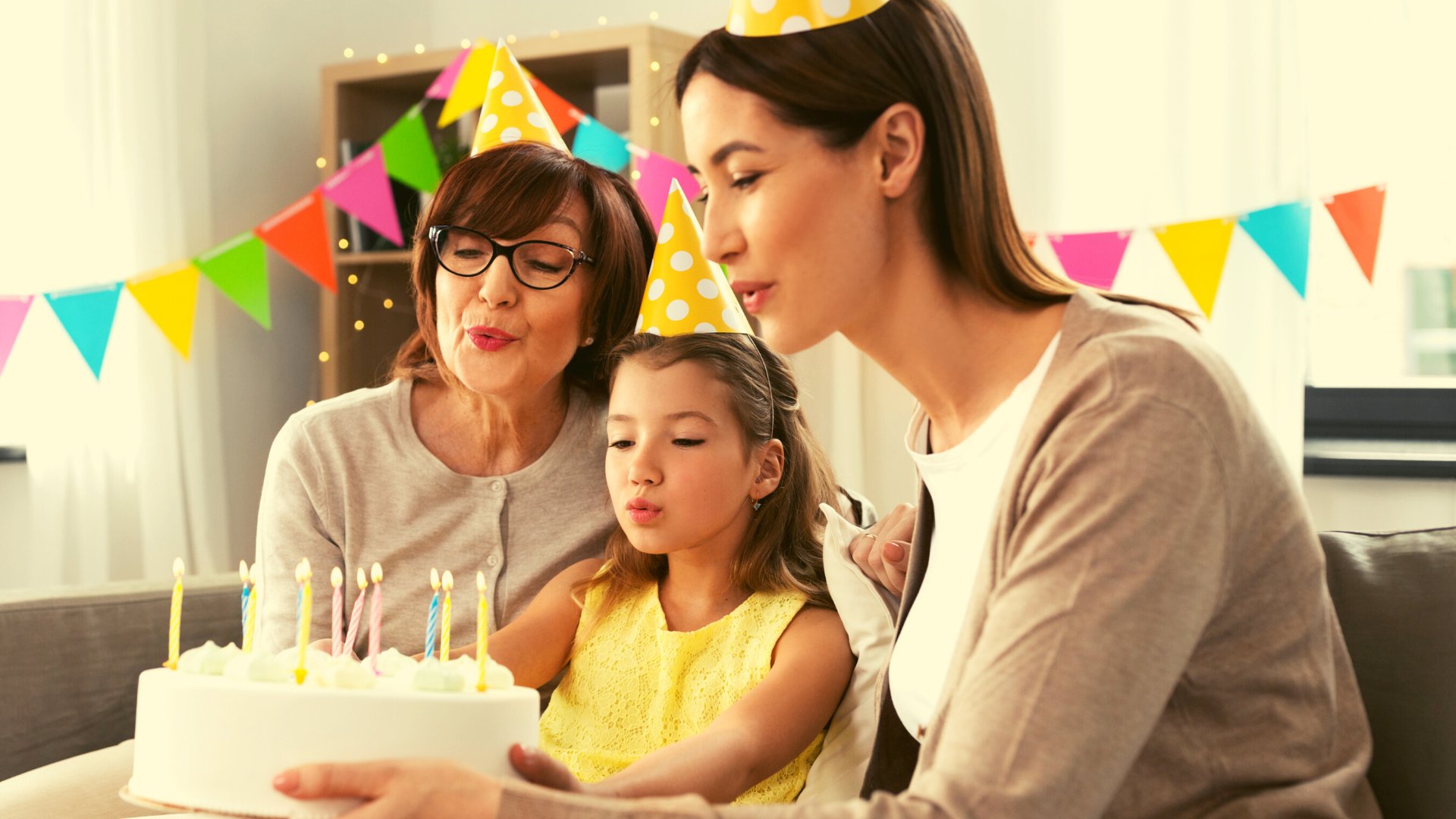 Avó, mãe e filha pequena sentadas em mesa com chapéus de festa, em frente à bolo com velinhas