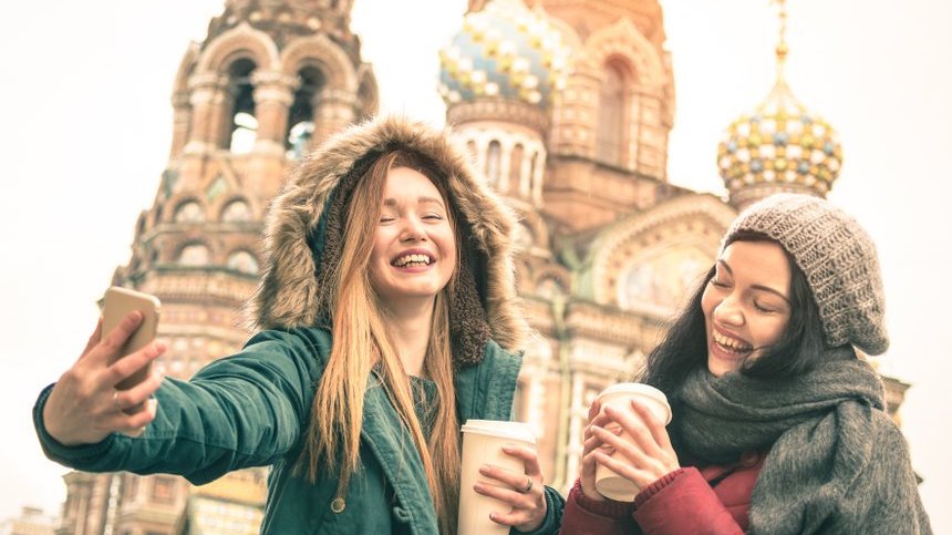 Viewapart / 123rf Duas amigas com café na mão tirando uma foto em frente a uma igreja