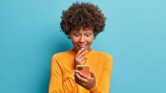 Cast Of Thousands / Shutterstock Mulher surpresa olhando para o celular que está em sua mão.