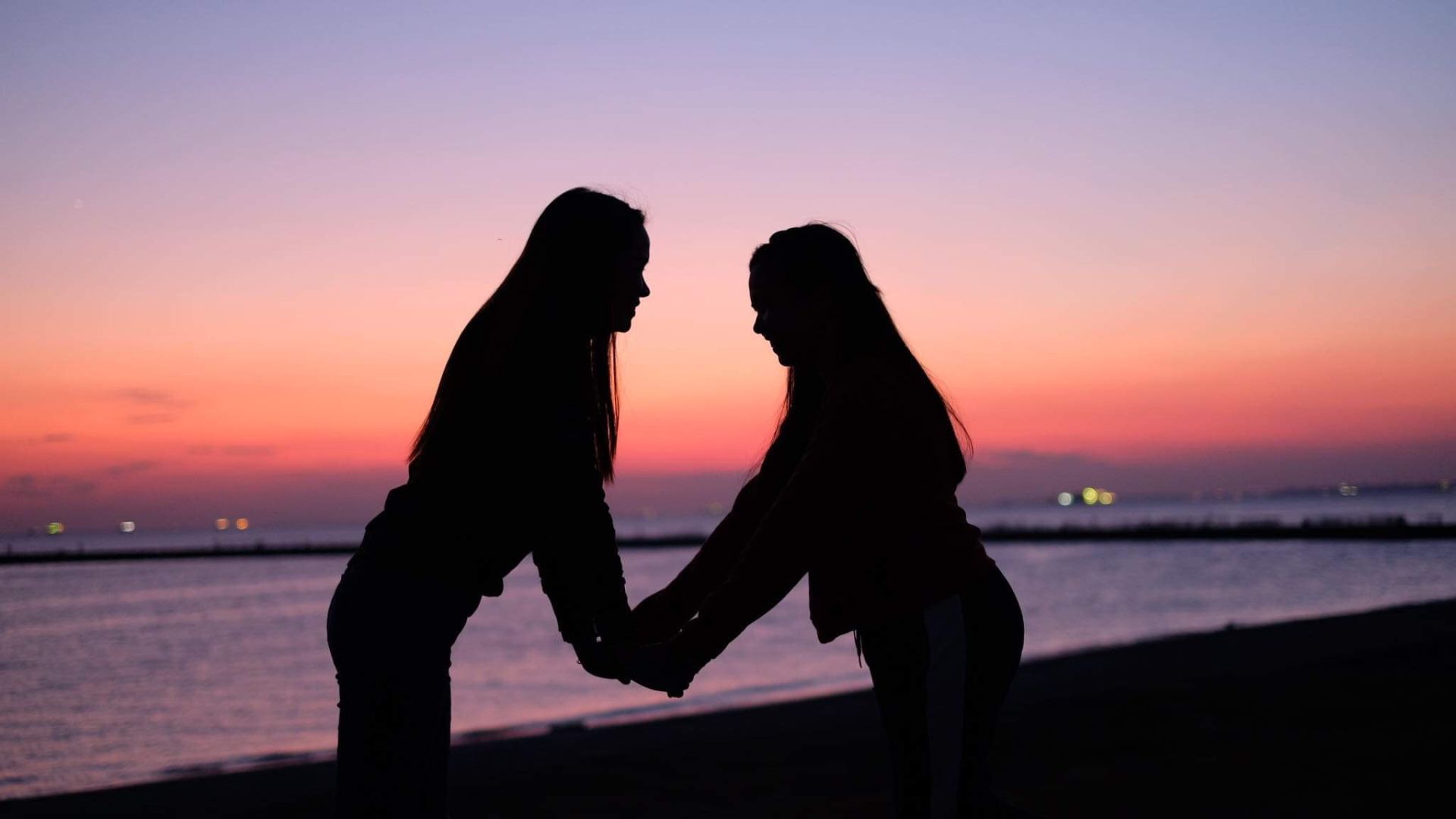 Imagem de duas amigas dando as mãos uma de frente para a outra na praia e ao fundo uma paisagem de céu alaranjado.