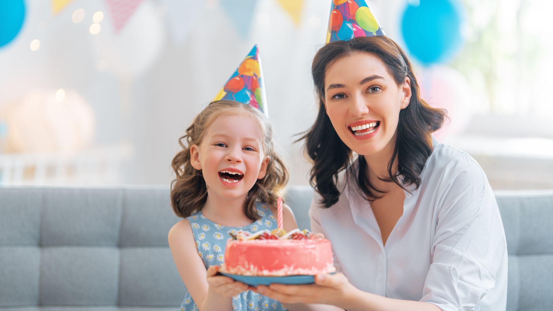Imagem de uma mulher ao lado de uma criança com chapéu de aniversário, elas estão segurando um bolo rosa de aniversário e bexigas atrás delas.
