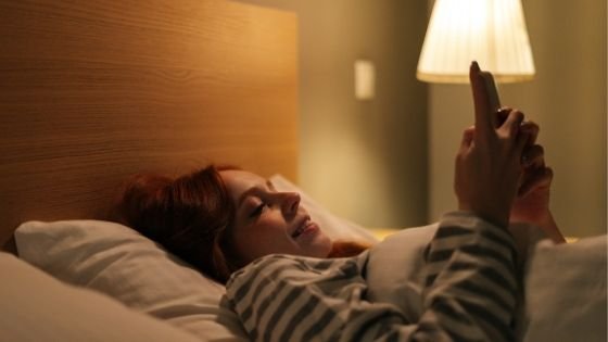 Mulher deitada na cama mexendo no celular com um abajur ligado atrás dela.