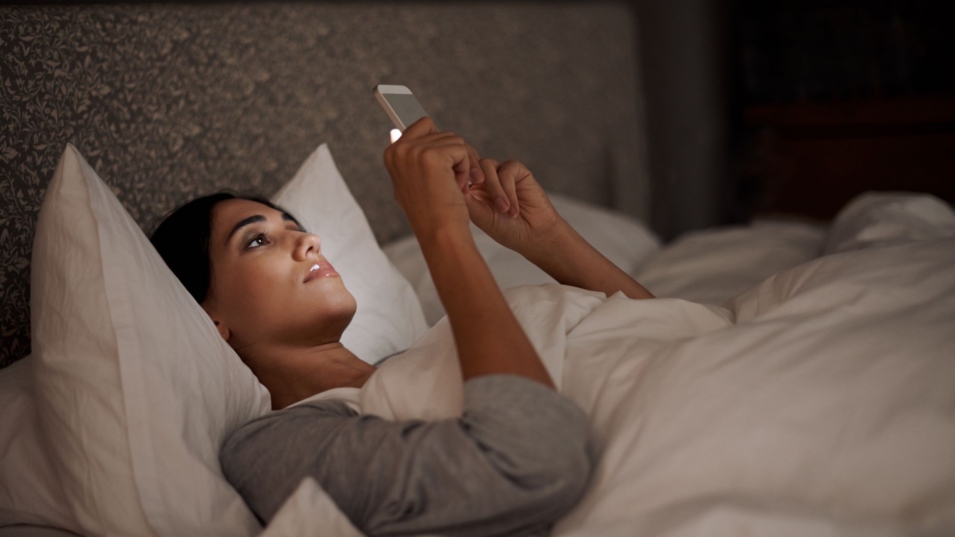 Imagem de uma mulher olhando o celular, deitada em sua cama antes de dormir