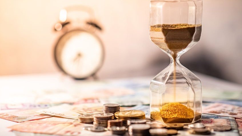 benjawansittidech / 123RF Ampulheta na mesa com notas e moedas e cronômetro de contagem regressiva ao fundo.