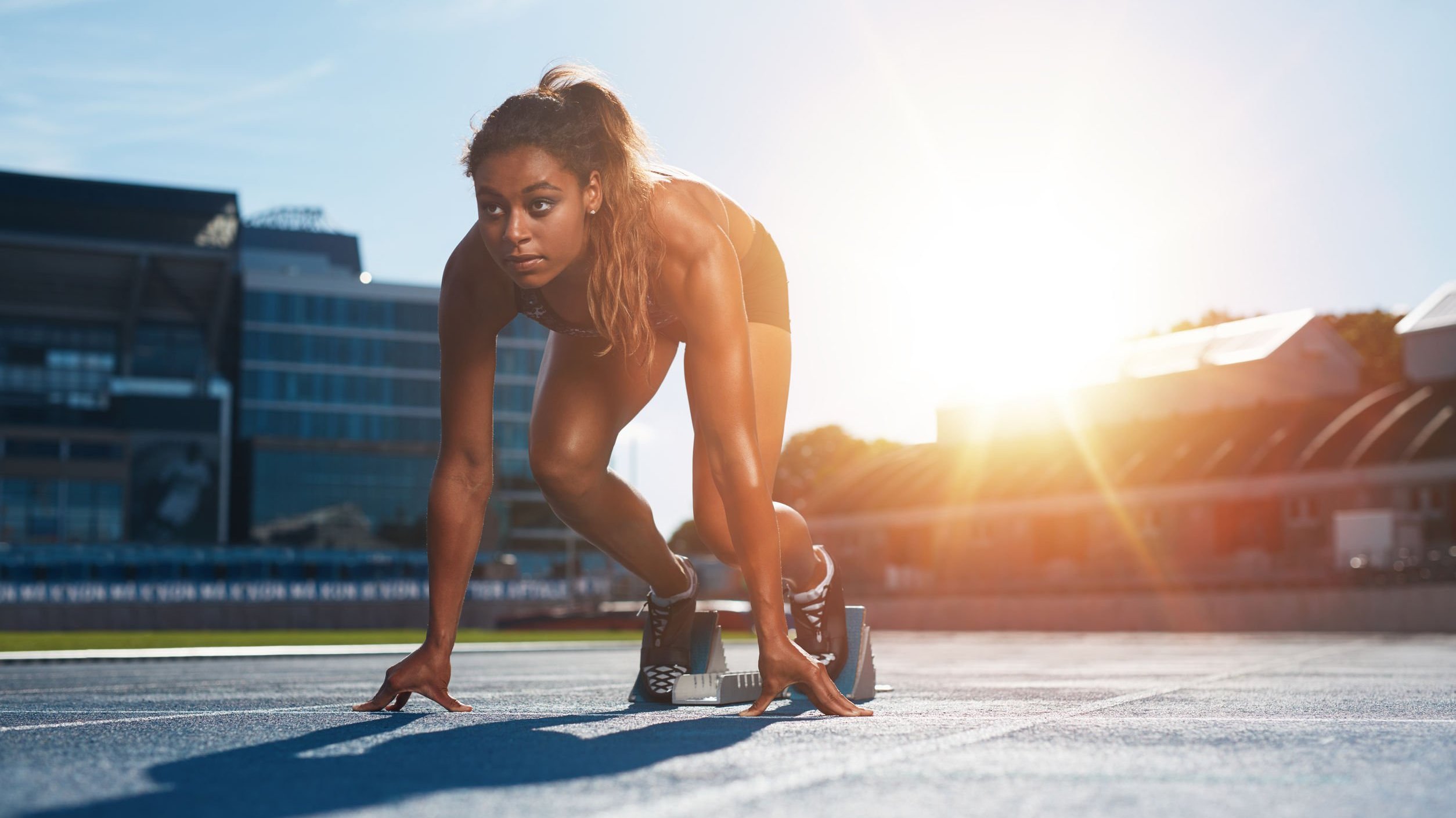 Atleta feminina, praticante de atletismo, em pista de corrida ao ar livre em dia ensolarado, em posição de largada.