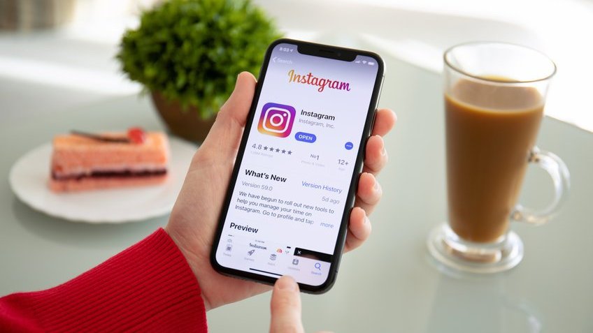 Perfil do instagram aberto no celular. Ao fundo, uma fatia de bolo, café e uma planta estão em cima da mesa