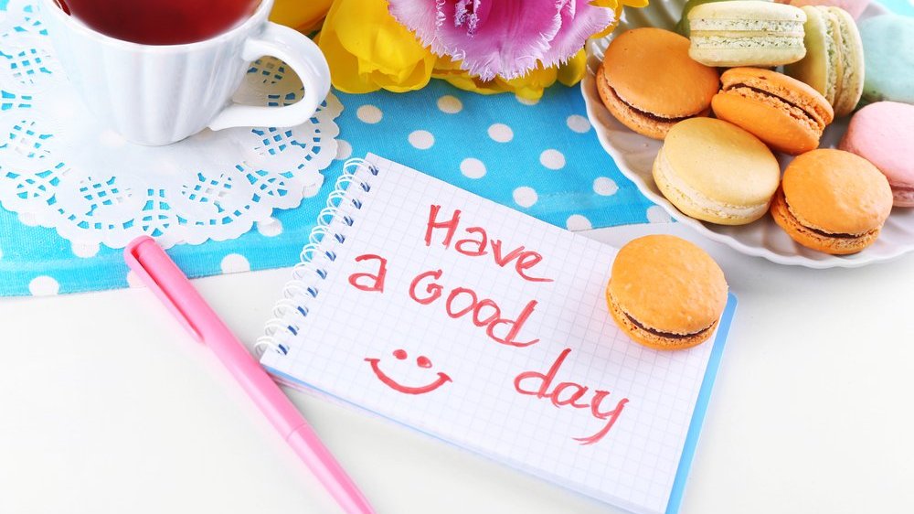 Recado em uma caderneta, escrito tenha um bom dia em inglês. Ao redor, uma caneta cor de rosa, uma xícara e um prato com macarons.