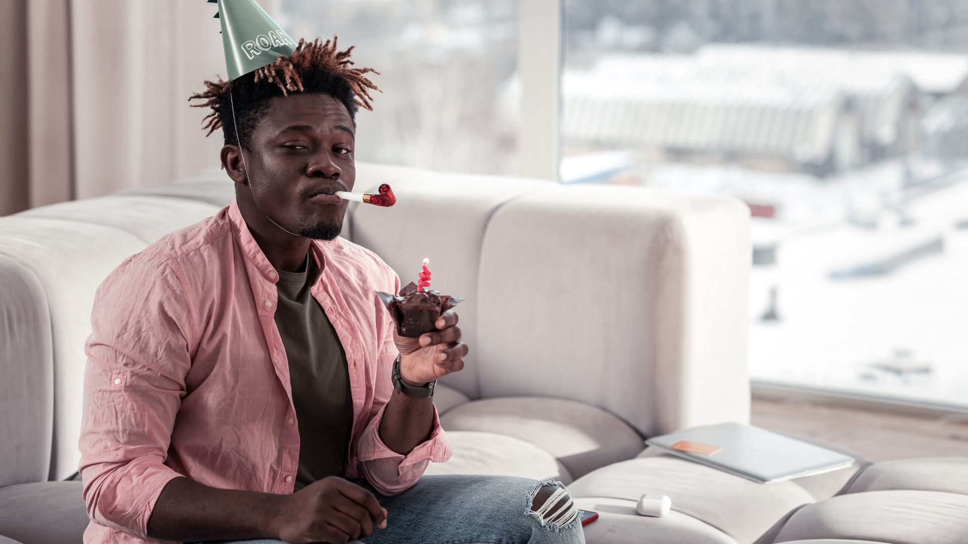 Imagem de um homem negro com chapéu de aniversário, com uma língua de sogra na boca e um cupcake com uma vela no topo, na mão sentado no sofá.