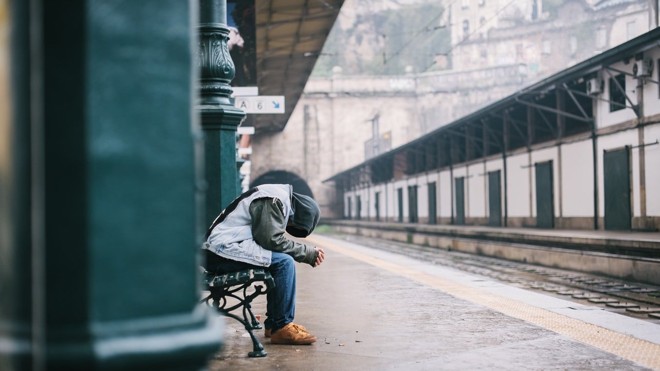 Pessoa vestindo capuz e sentada em estação de trem, segurando as duas mãos com a cabeça baixa.