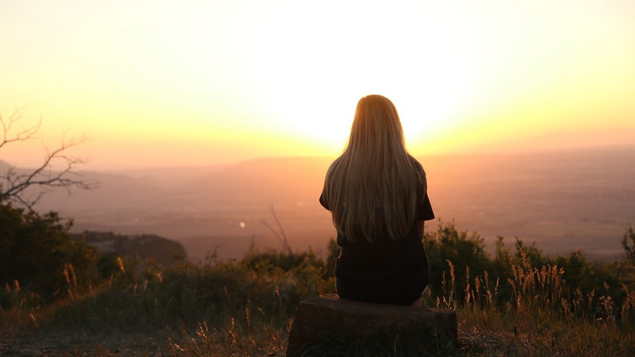 Mulher sentada no topo de uma montanha, de costas, observando o pôr do sol.