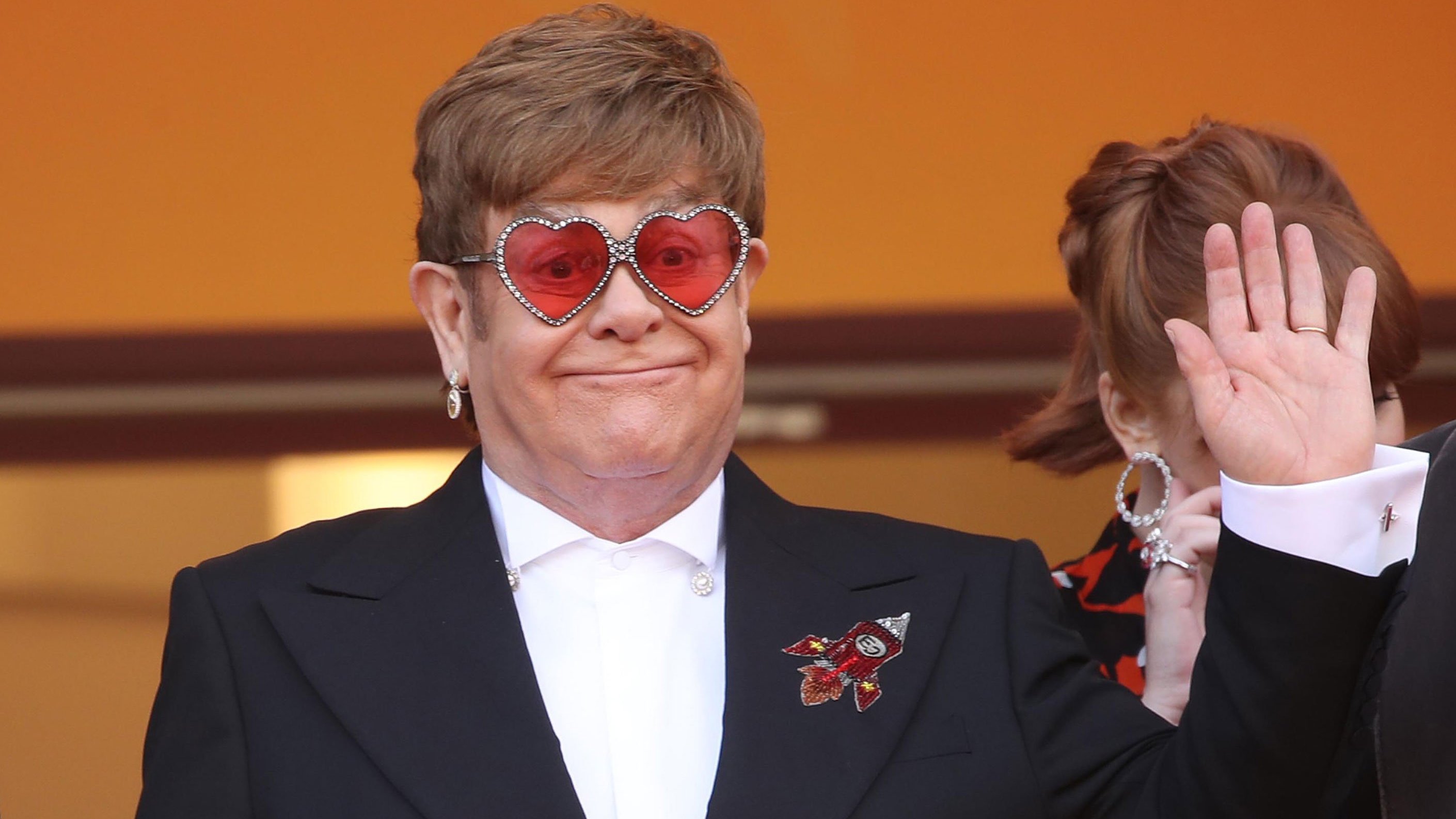 Elton John acenando em evento, usando terno preto com detalhe de flor, e óculos vernelhos em formato de coração, com brilhantes na armação