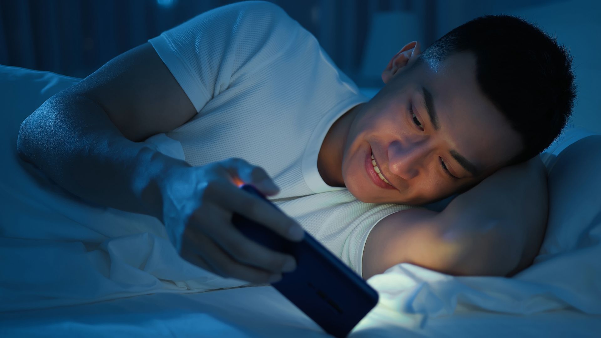 Imagem de um homem deitado na cama olhando para o celular e sorrindo
