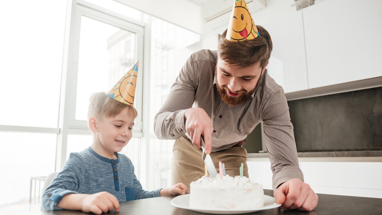 Menino e homem brancos com chapéu de aniversário enquanto cortam um bolo.