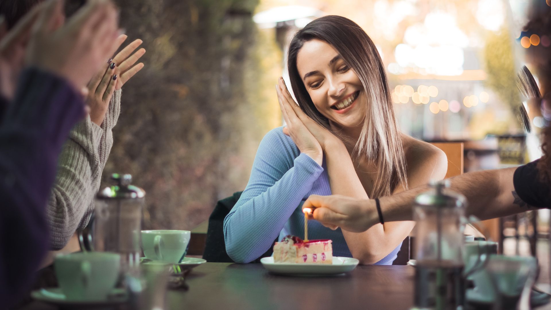 Imagem de uma moça recebendo uma festa de aniversário e sorrindo na frente de um pedaço de bolo com uma vela no topo