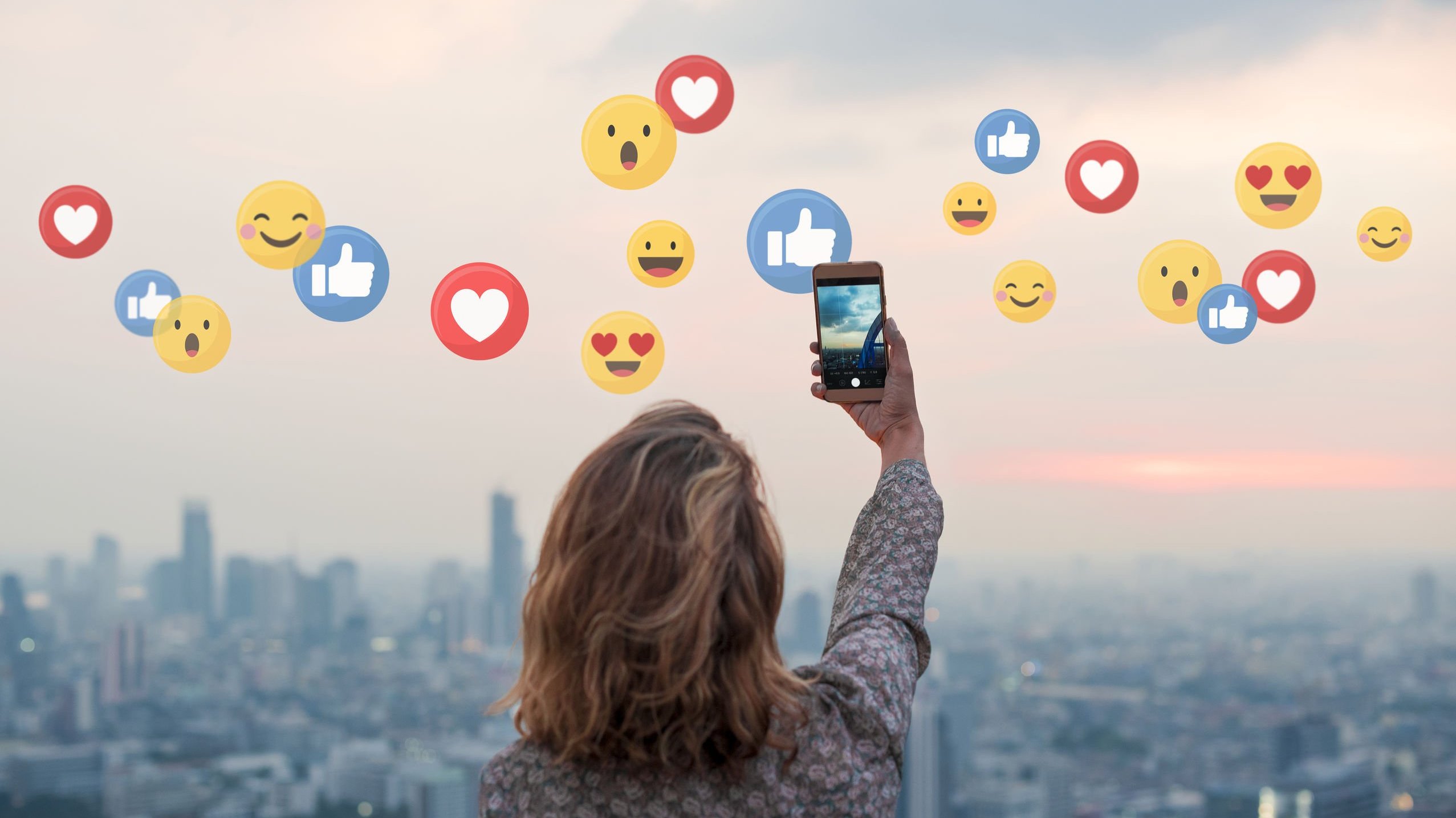 Mulher tirando selfie em meio à paisagem urbana com pôr do sol e vários emojis de reações do Facebook.