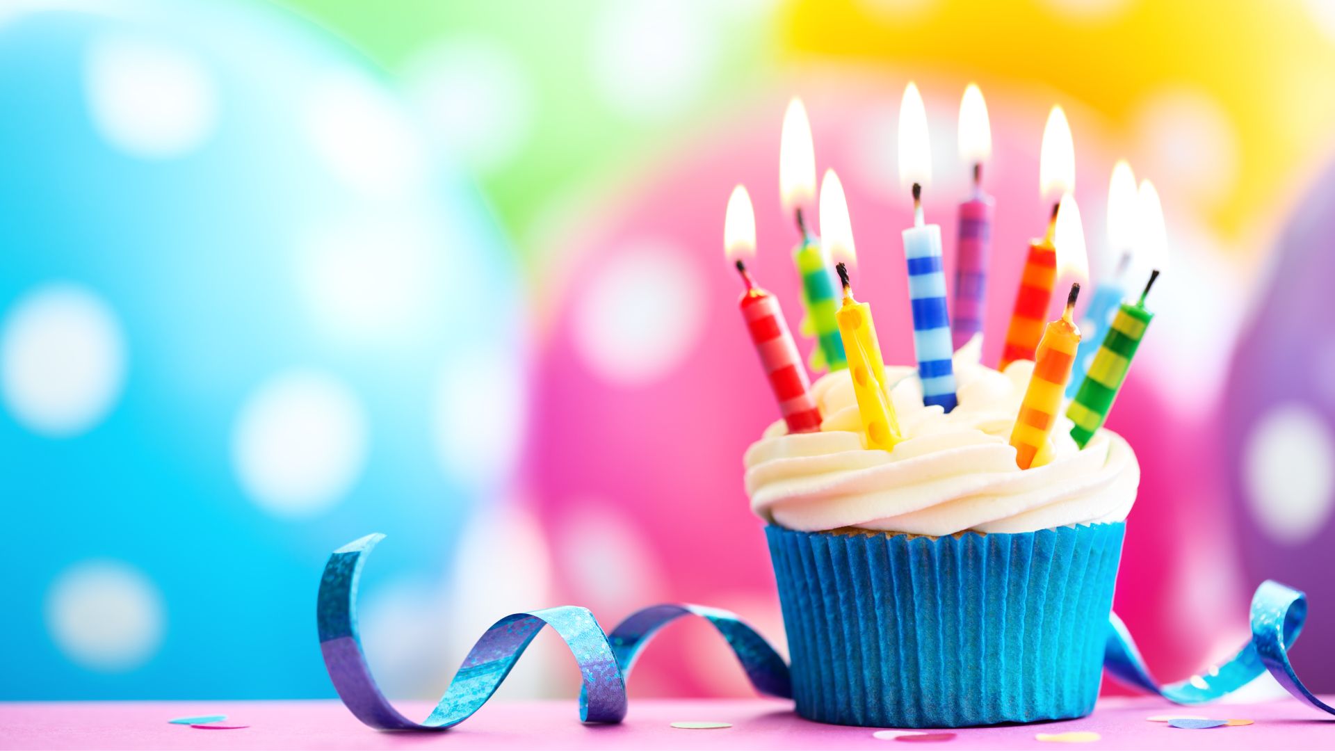 Imagem de um cupcake com velas coloridas em cima, uma fita de presente e balões coloridos desfocados no fundo