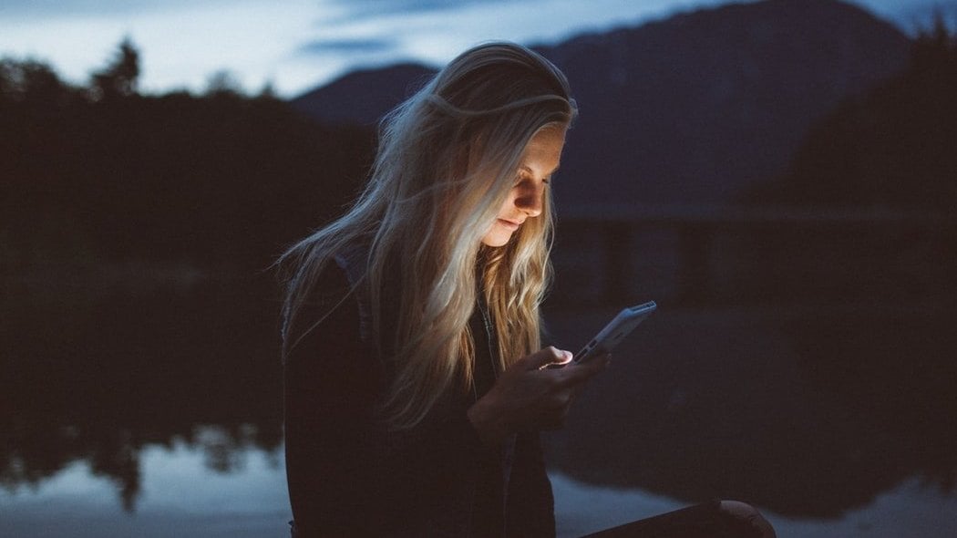 Mulher sentada próxima a um lago no começo da noite, mexendo no celular.