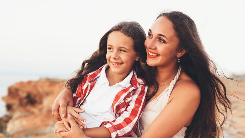 Mulher e menina brancas sorrindo abraçadas.