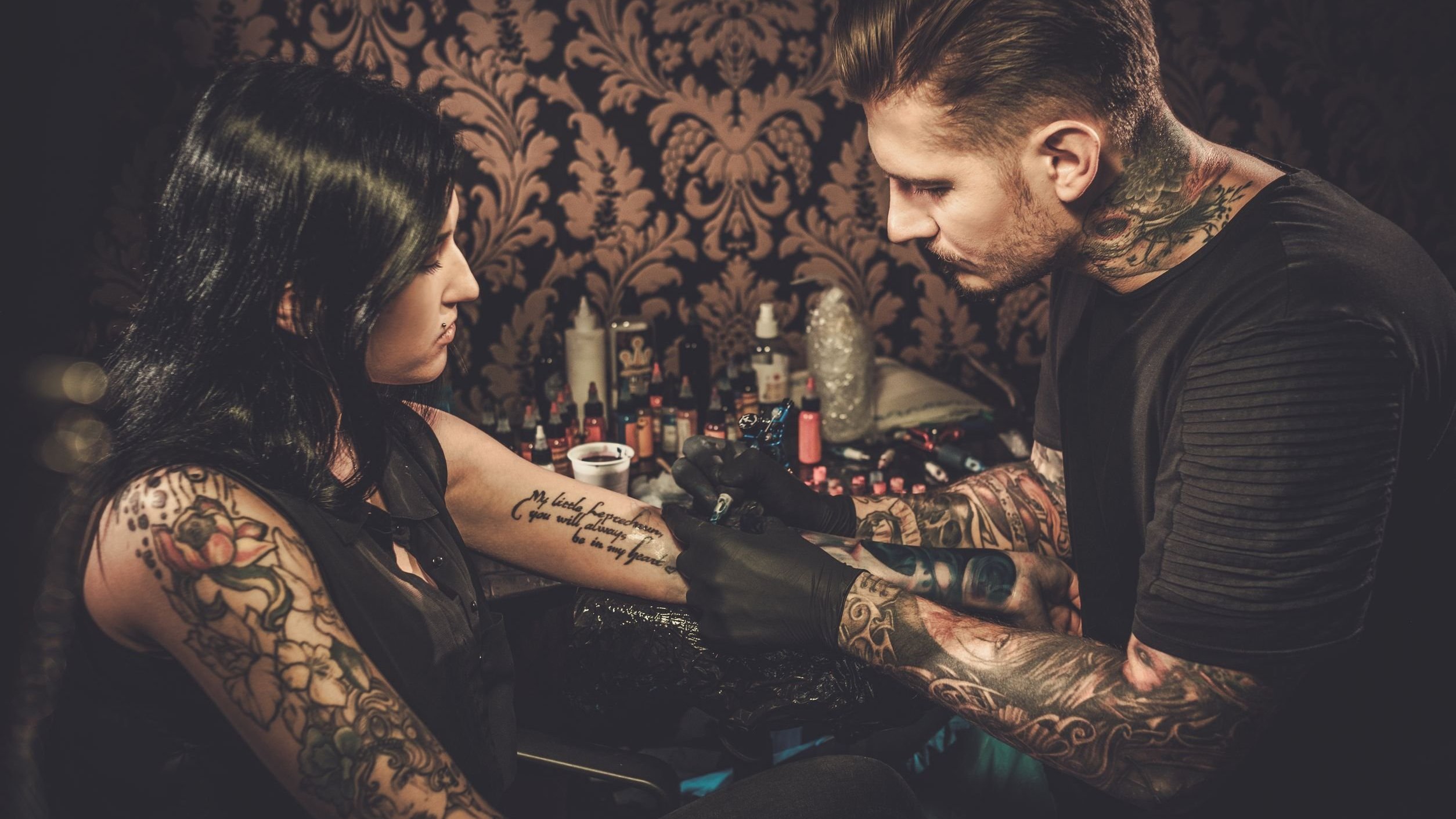 Homem e mulher cobertos por tatuagens. O homem está tatuando o braço esquerdo da mulher.