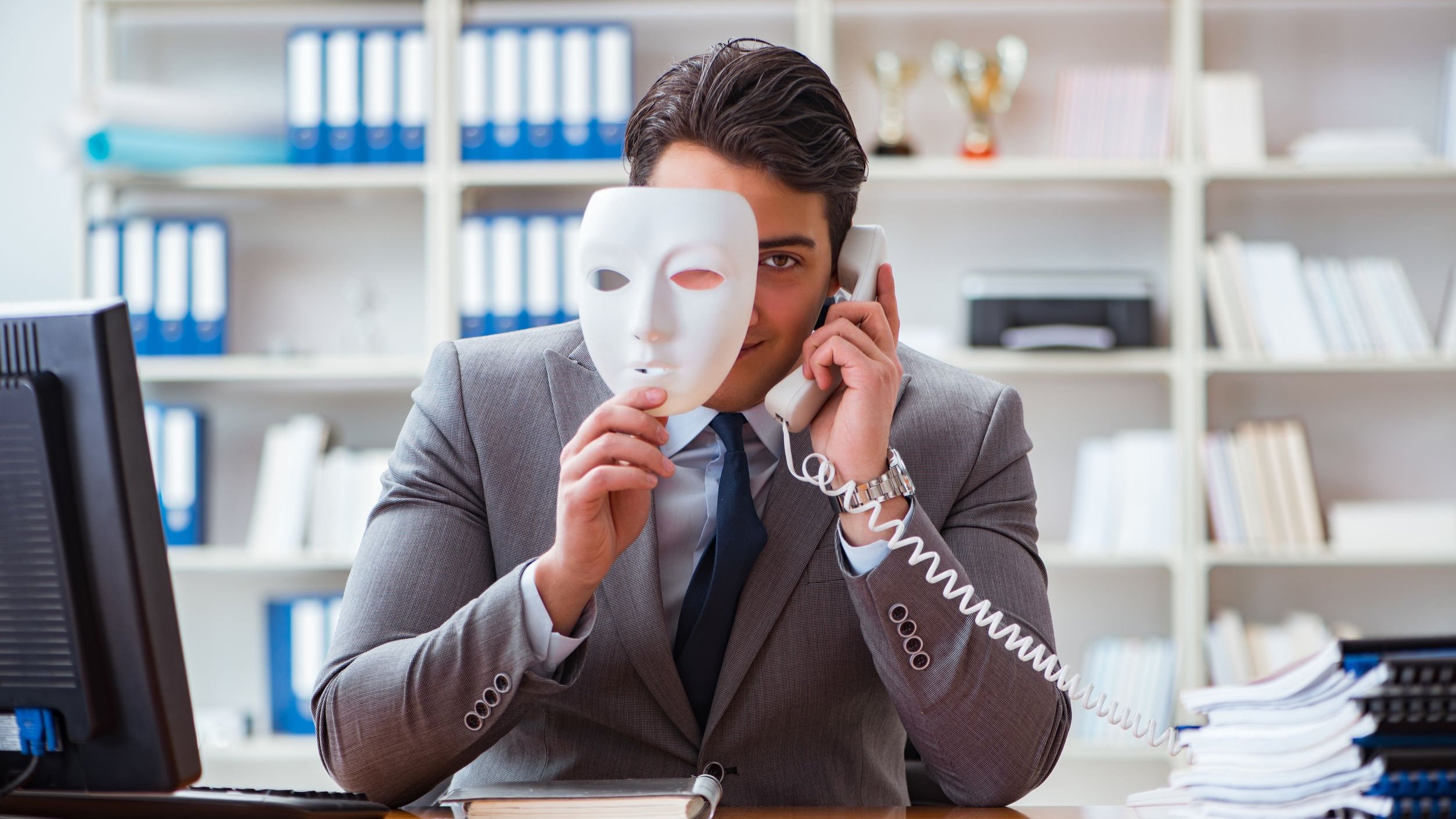 Homem em escritório, usando o telefone, segura uma máscara branca em frente ao seu rosto.