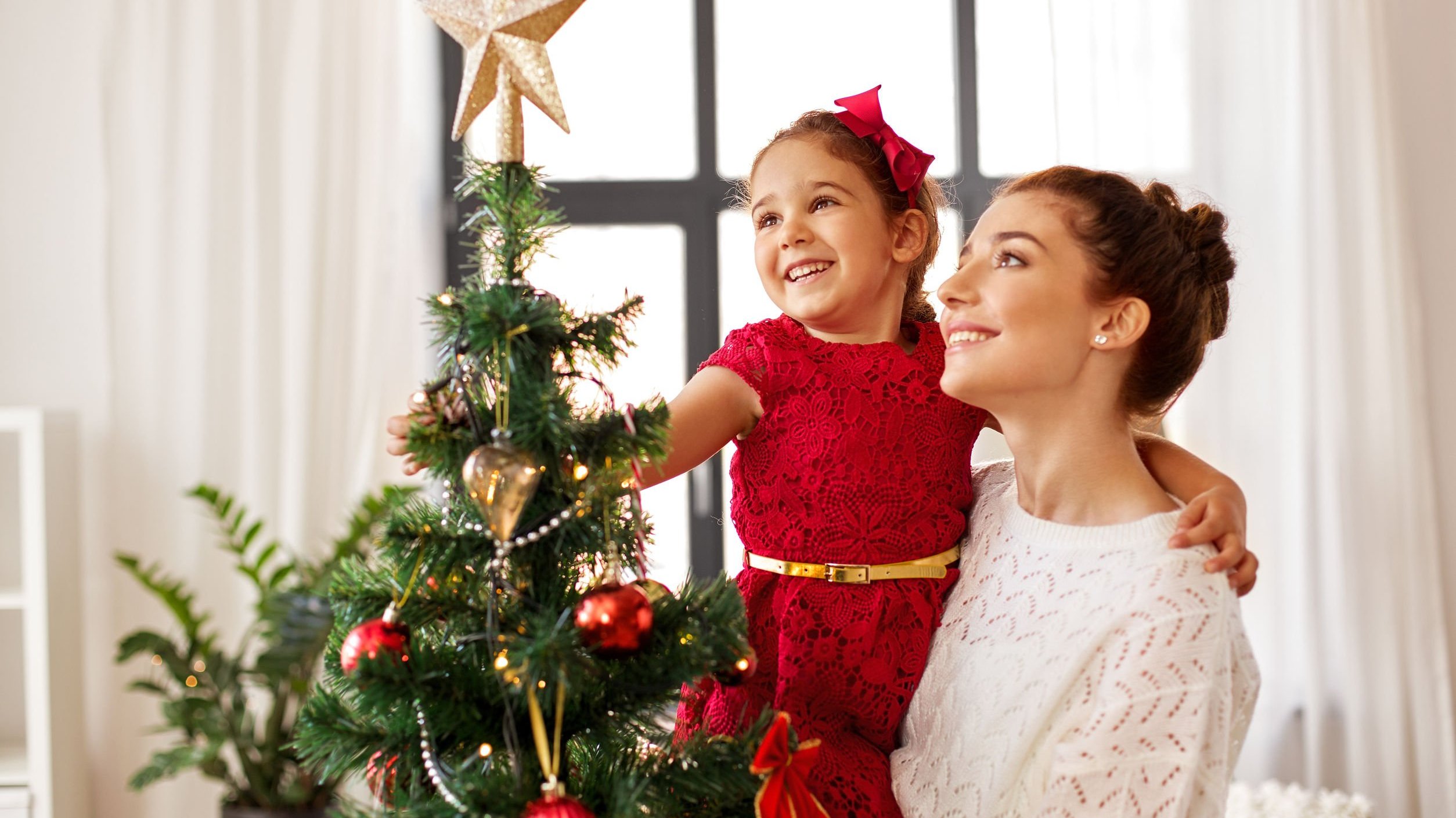 Mulher segurando menina no colo em frente de árvore de Natal. A menina está colocando uma estrela no topo da árvore.