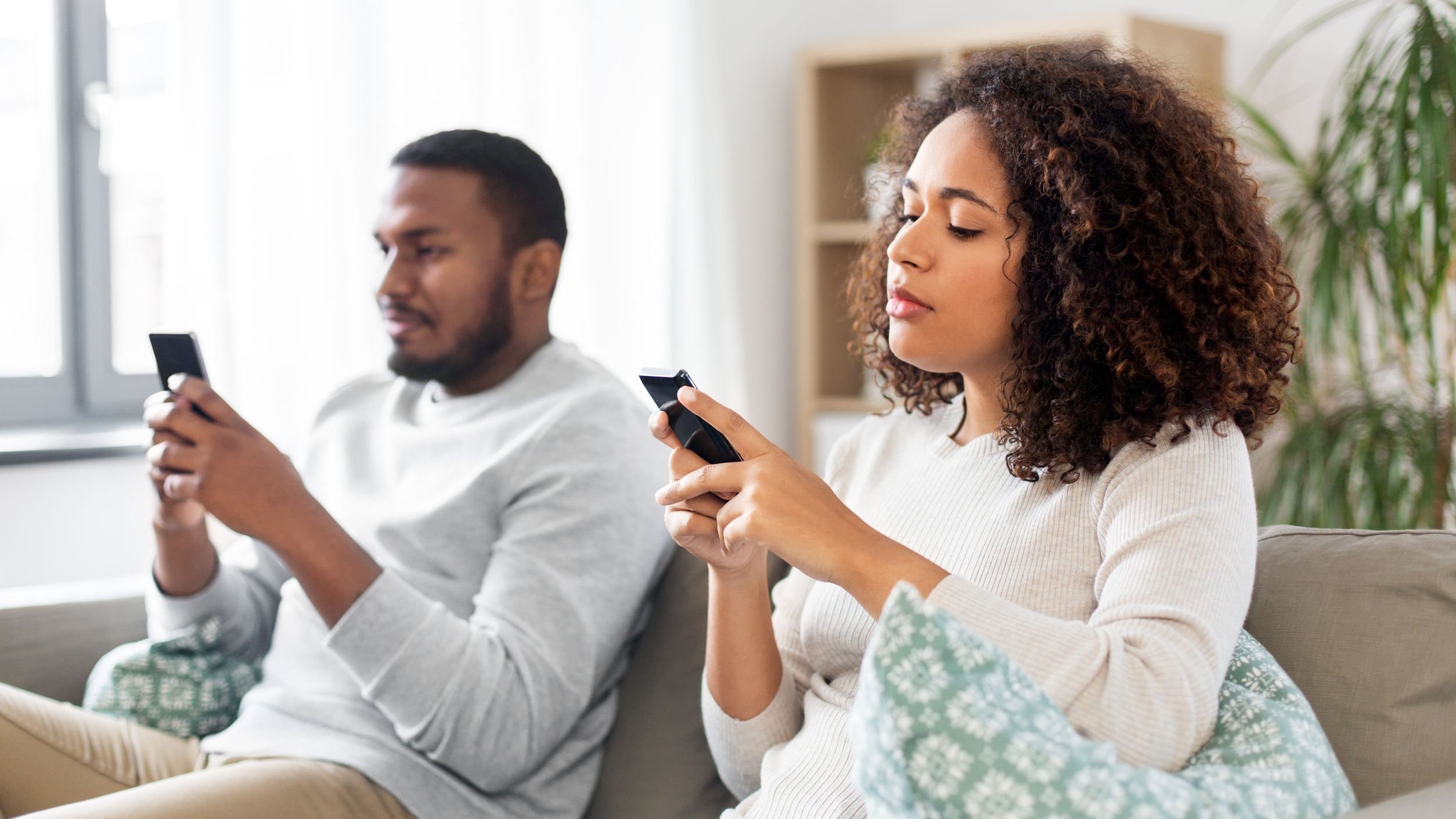 Homem e mulher sentados em sofá, ambos mexendo em seus celulares.