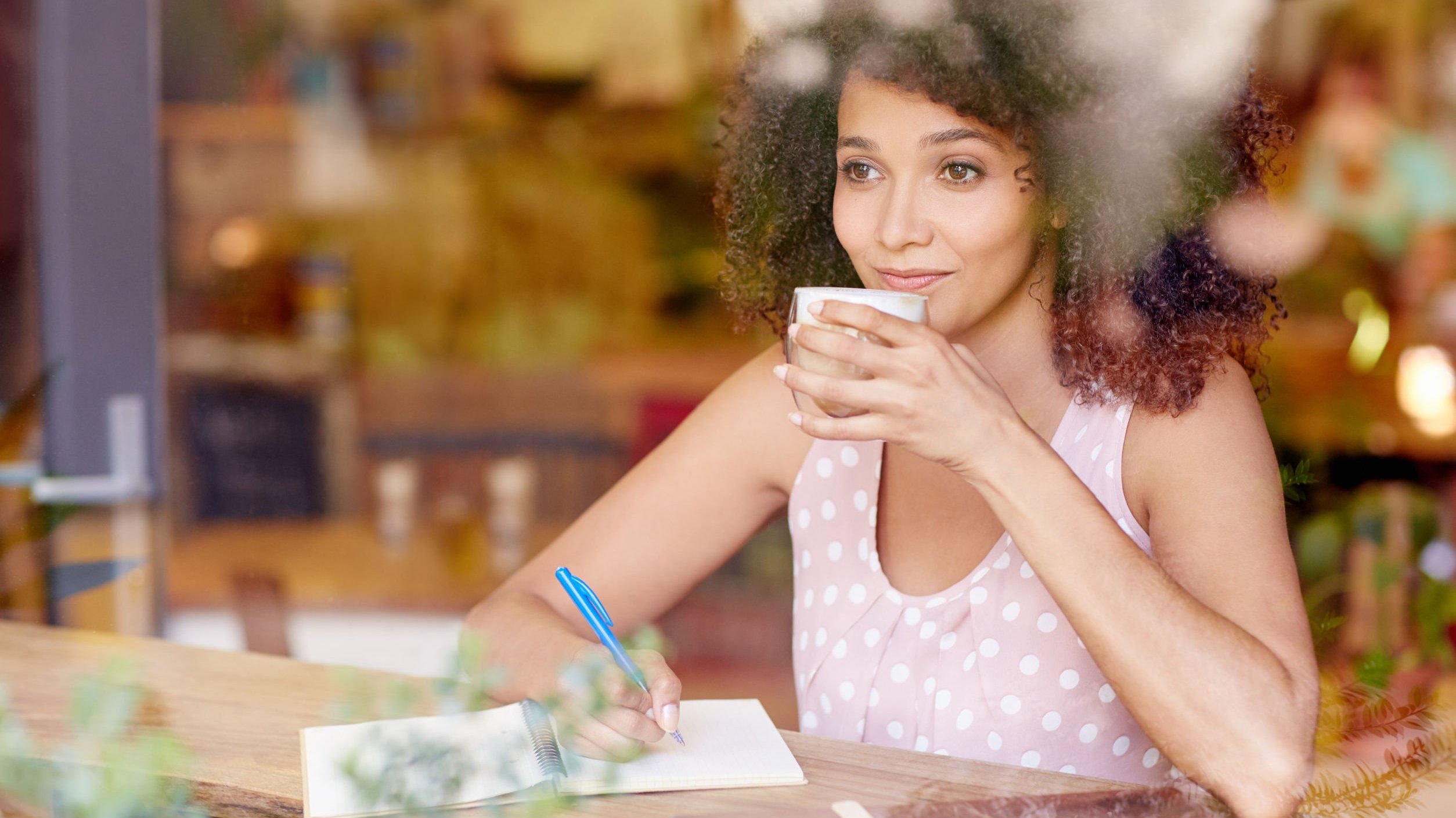 Mulher sentada em uma mesa, escrevendo em um caderno e bebendo café