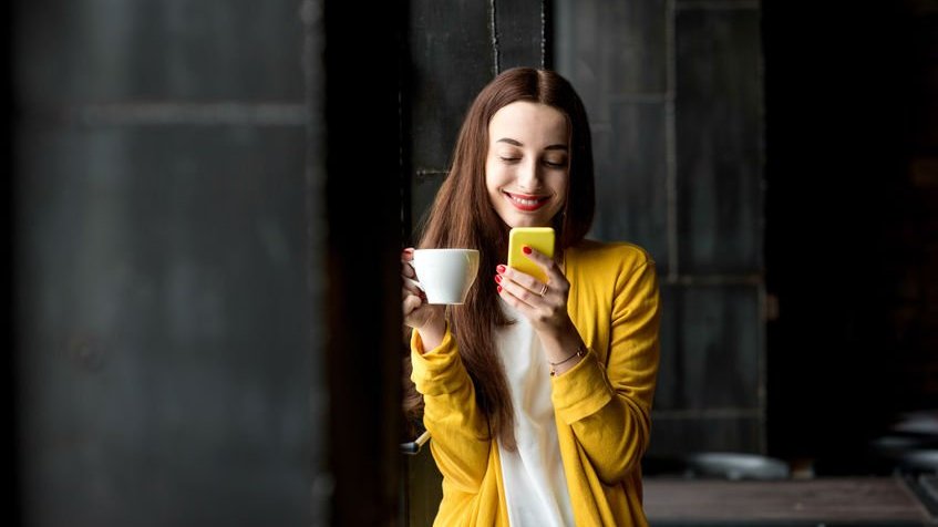Mulher olhando para celular segurando xícara na outra mão sorrindo