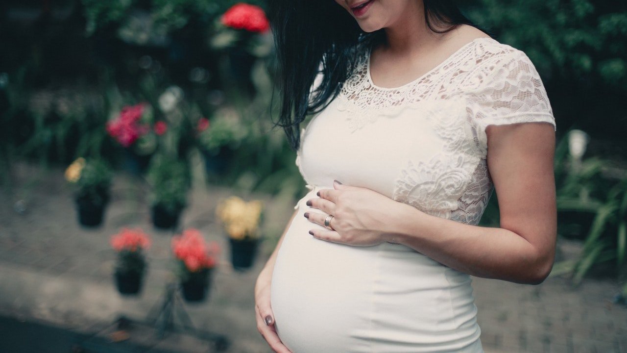 Mulher grávida usando vestido branco segura sua barriga com as duas mãos.