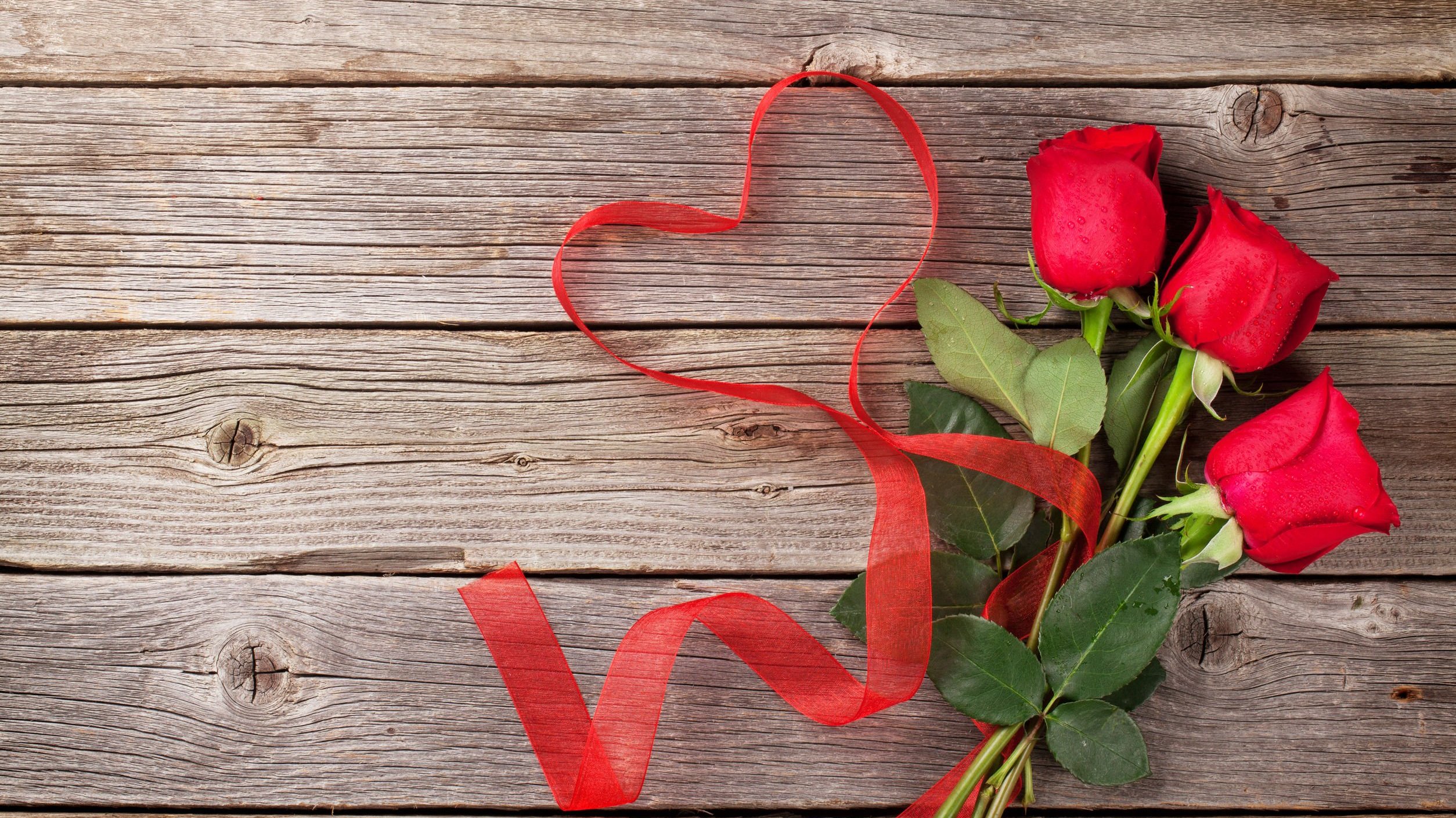 Rosas vermelhas ao lado de fita posta em formato de coração. Os dois estão sobre a madeira