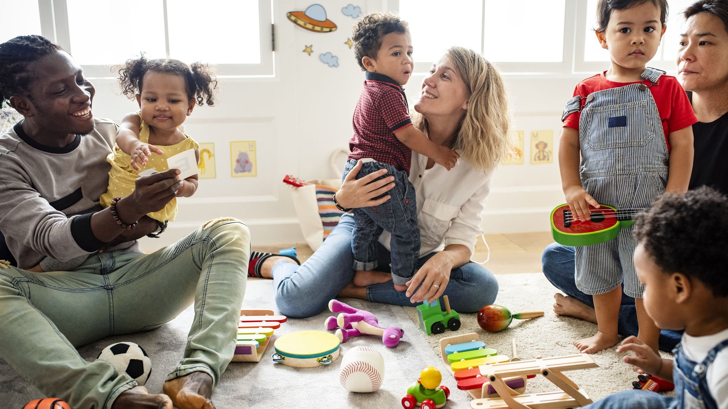 Crianças pequenas com seus pais, em uma sala com brinquedos espalhados pelo chão.