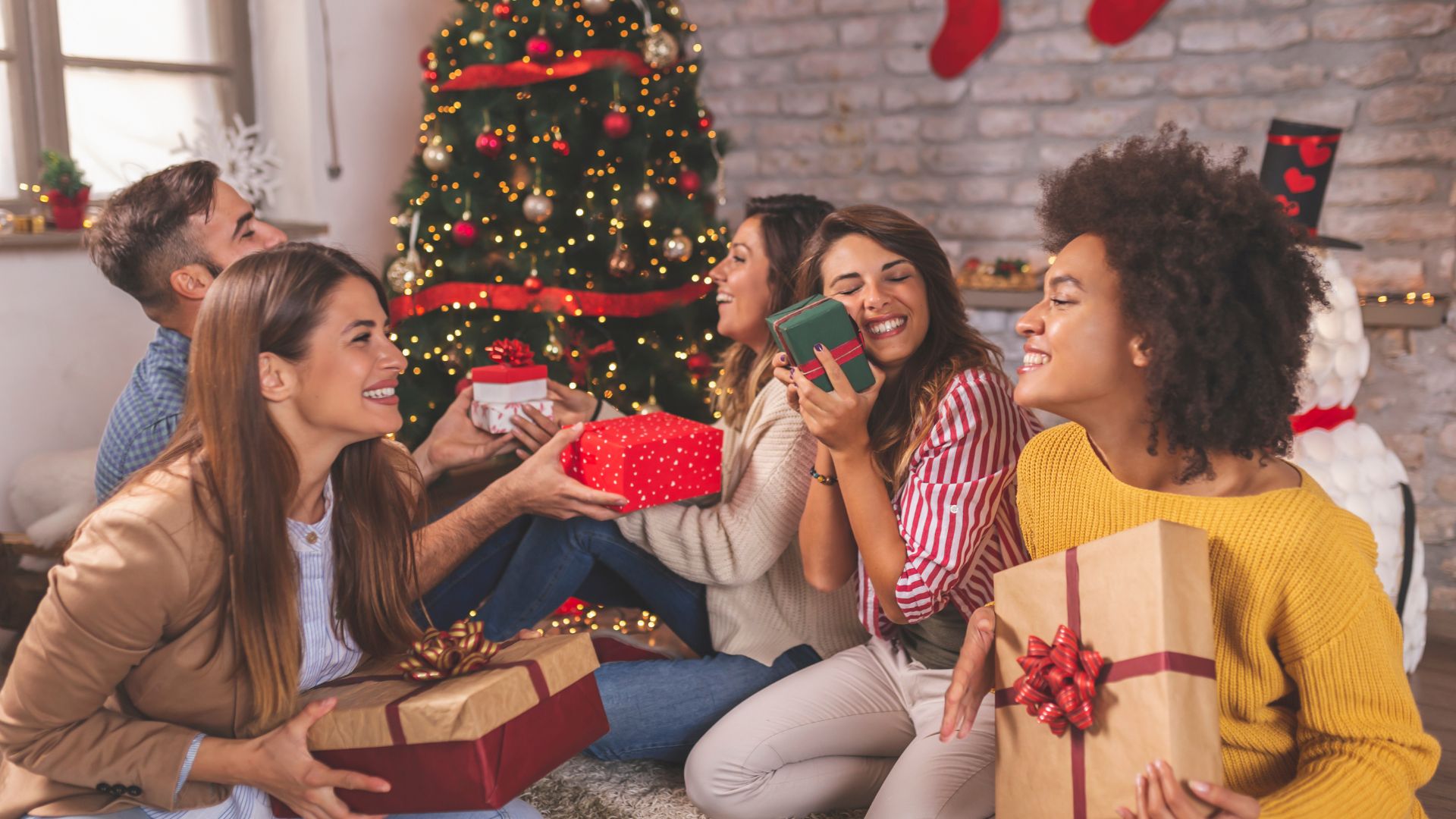 Imagem de várias pessoas felizes e sorridentes sentadas no chão trocando presentes de Natal. Ao fundo uma linda árvore decorada com enfeites natalinos.