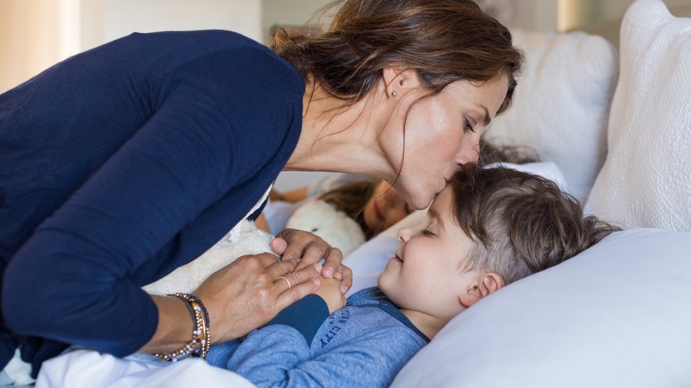 Mãe dando um beijo de boa noite na testa do filho adormecido.