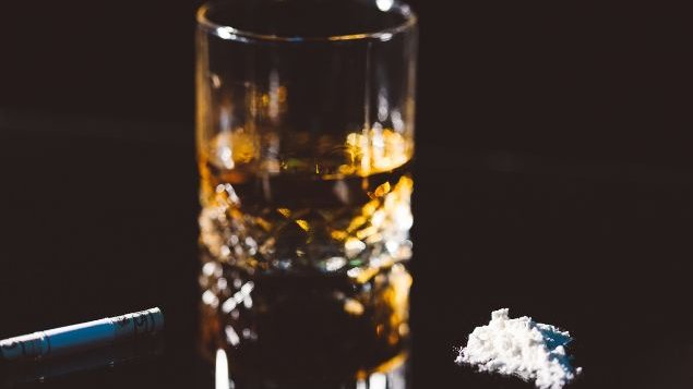 Imagem de um copo com cerveja e, ao lado, um cigarro aceso e drogas