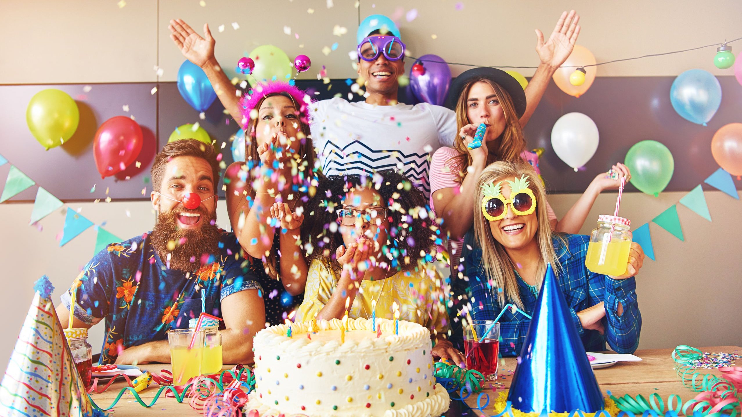 Grupo com seis pessoas usando máscaras e acessórios de festa comemora atrás de um bolo de aniversário.