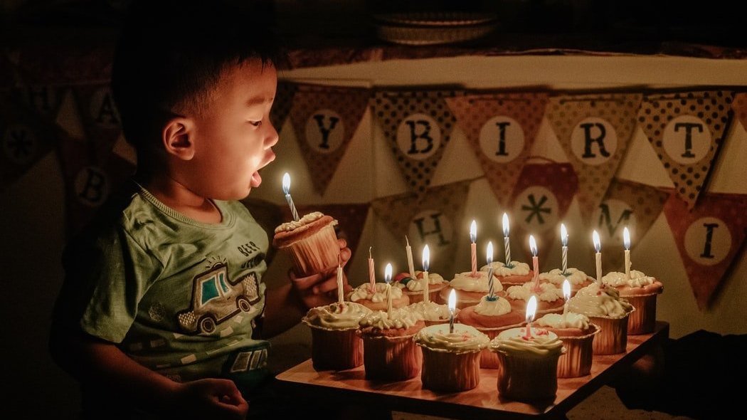 Menino em frente de muitos cupcakes, com uma vela no topo de cada. Ele segura um dos cupcakes com a vela acesa, e o leva em direção a sua boca.