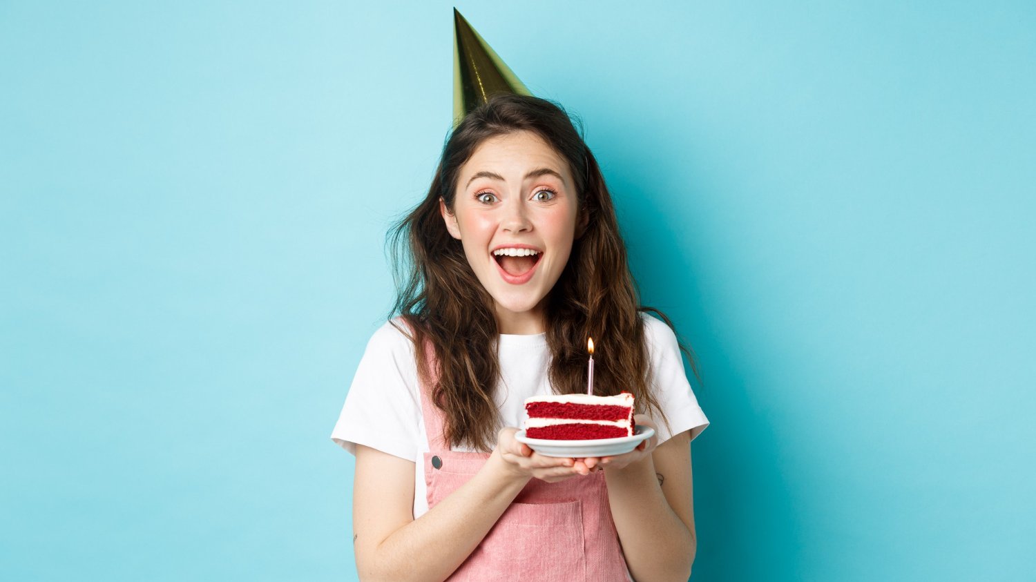 Mulher branca de cabelos grandes e escuros sorrindo enquanto segura um pedaço de bolo com uma vela em cima.