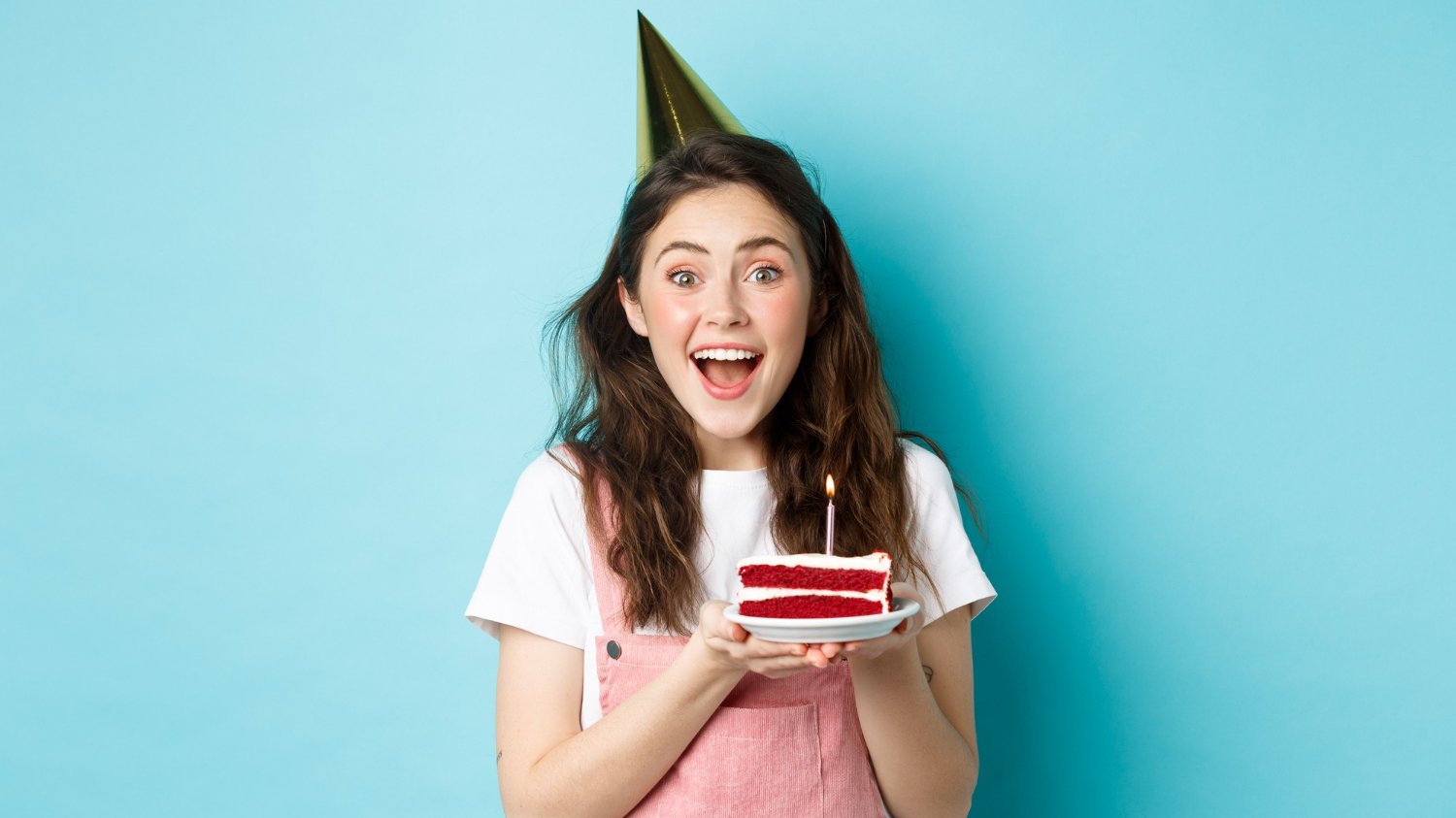 enzoix / Freepik Mulher branca de cabelos grandes e escuros sorrindo enquanto segura um pedaço de bolo com uma vela em cima.