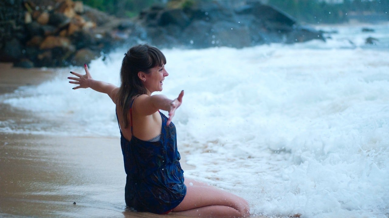 Mulher de maiô ajoelhada na água do mar na praia, com os braços abertos.