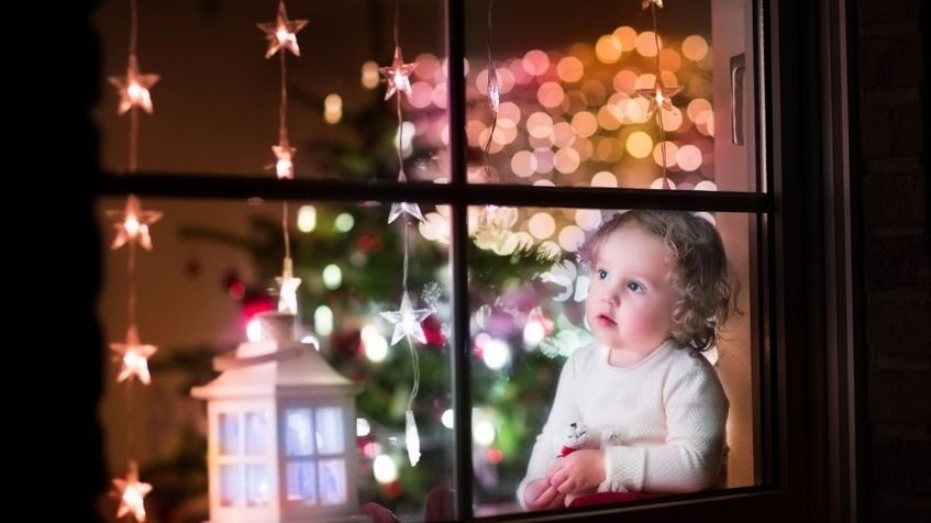 Menina sentada com um urso de brinquedo em casa durante o Natal vista de fora para uma sala de jantar decorada com árvore e luzes